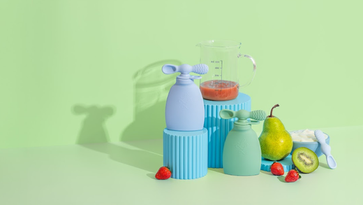 Two colorful reusable pouches displayed with fresh strawberries, pear, kiwi, yogurt, and a glass jug of smoothie on a pastel green background.