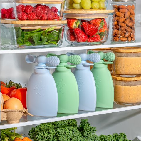 Reusable pouches in pastel colors neatly arranged in a fridge surrounded by fresh fruits, vegetables, nuts, and smoothies.