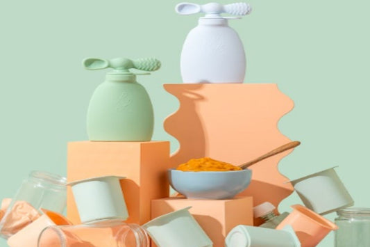 Green and white Palmetto Pouches displayed on pastel blocks beside a bowl of puree and scattered disposable containers—emphasizing reusable, sustainable baby feeding products
