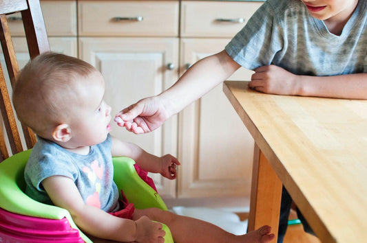 Baby sits in a booster seat, experiencing one of the first foods as big brother introduces solids with a yogurt bite—a key milestone in determining when babies can eat baby food.