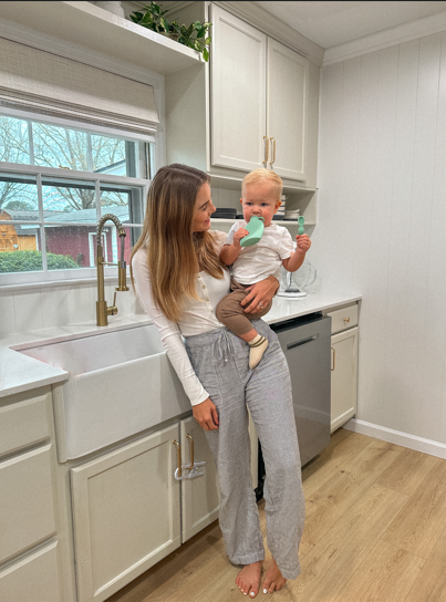 A mother holding her baby in the kitchen while the baby enjoys a homemade puree from a reusable food pouch, promoting healthy eating habits.