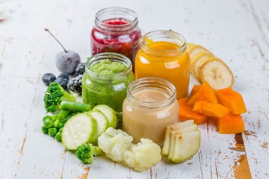 Assorted jars of homemade baby food with ingredients on display, including pureed greens, carrots, apples, and bananas, ideal for babies starting on solids.