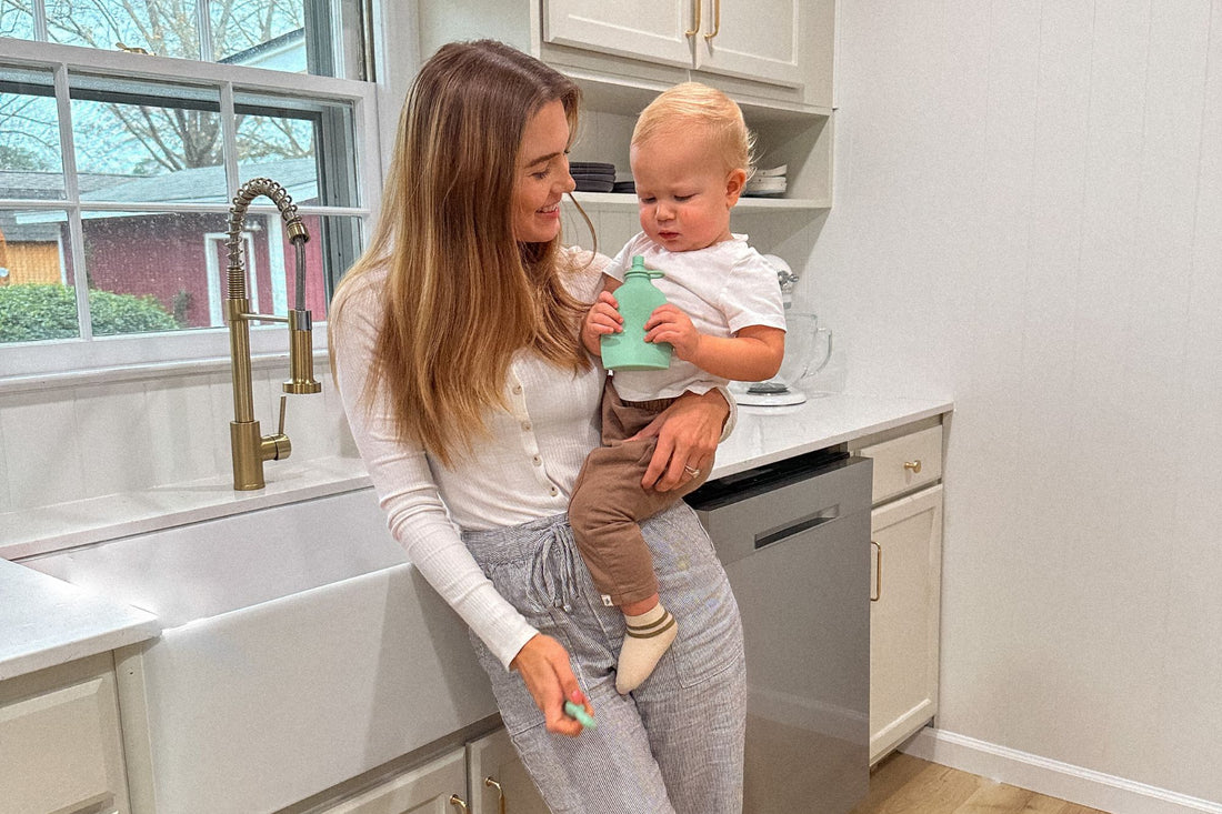 A mother holding her baby in a kitchen, while the baby drinks from a reusable Palmetto Pouch by Lowcountry Littles. The pouch’s soft silicone design is perfect for both babies and toddlers, offering a mess-free snack solution.